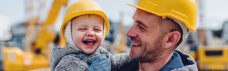 Ein lachendes Kleinkind mit Bauhelm sitzt auf dem Arm seines Vaters, der ebenfalls einen Helm trägt – beide stehen auf einer Baustelle. Das Bild symbolisiert, wie die RÖDL BAU.GRUPPE berufliches Engagement und Familienleben miteinander verbindet.